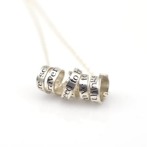 May include: Silver necklace with a spiral of rings that have words engraved on them. The words are "be braver", "need to be", "have to", and "things".