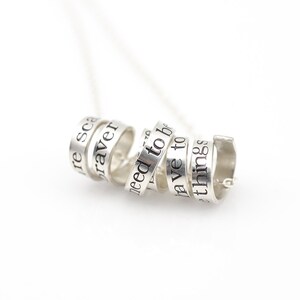 May include: Silver necklace with multiple rings that have words engraved on them. The words are "we need to be braver", "we have to be", and "things are scary".