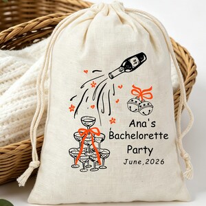May include: A cream-colored drawstring fabric pouch featuring a black and orange graphic of a champagne bottle pouring into a tower of glasses, two jingle bells with an orange bow, and the text Ana's Bachelorette Party June, 2026.