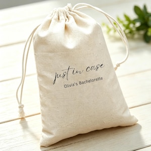 May include: A natural beige cotton drawstring pouch featuring the black printed text "just in case" in a cursive script font, followed by the text "Olivia's Bachelorette" in a clean, sans-serif typeface centered on the front of the bag.