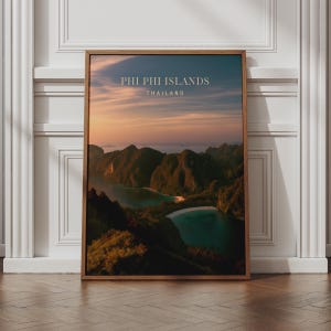May include: A framed vertical wall art print featuring a sunset landscape of the Phi Phi Islands in Thailand. The image displays dark green mountains surrounding turquoise bays under a soft orange and purple sky with the text PHI PHI ISLANDS THAILAND.