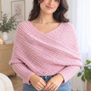 May include: A light pink knitted wrap sweater featuring a textured stitch pattern, long sleeves with ribbed cuffs, and a V-neckline created by overlapping fabric panels that cross over the chest, paired with blue denim jeans.