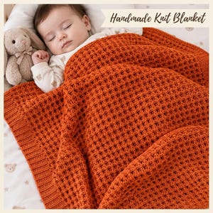 May include: A burnt orange handmade knit blanket with a textured waffle stitch pattern and a ribbed border. The blanket is draped over a sleeping infant wearing a white long-sleeve shirt with bear prints, positioned next to a plush tan rabbit toy.