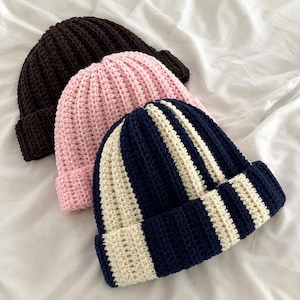May include: Three cuffed beanies made of crocheted yarn. One beanie is solid dark brown, one is solid light pink, and the third features vertical stripes in alternating navy blue and cream colors. Each hat has a thick, folded brim at the base.