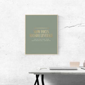May include: A rectangular wall art print featuring a sage green background with gold text that reads SLOW FOCUS. MASSIVE LEVERAGE. ARCHITECTED FOR EXPONENTIAL GROWTH. The print is framed in a thin gold border and displayed on a textured white wall.
