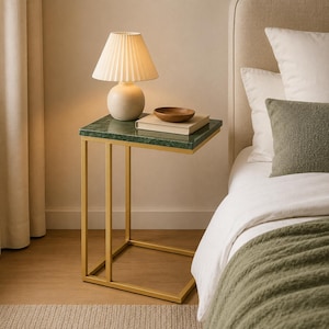 May include: A C-shaped side table with a dark green marble top and a gold-toned metal frame. The table holds a cream-colored lamp with a pleated shade, a small wooden bowl, and a book. The table is positioned next to a bed with pillows.