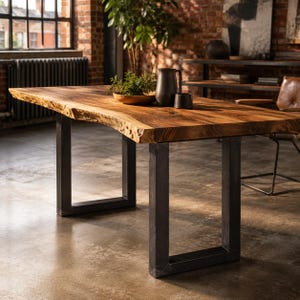 May include: A rectangular dining table featuring a thick, natural live-edge wood slab top with a warm brown grain pattern. The table is supported by two sturdy, matte black metal U-shaped legs. A small green plant and two dark ceramic vessels sit on top.