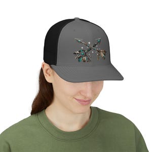 May include: A grey and black trucker hat featuring a graphic print of two crossed arrows adorned with turquoise stones, beads, and feathers. The design includes small white star accents and a detailed pattern on the arrow shafts and fletching.