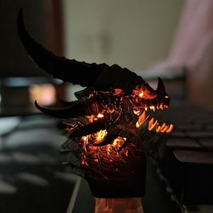 Pode incluir: Uma tecla de teclado em forma de cabeça de dragão escura e texturizada, com um chifre curvo proeminente e dentes afiados. A escultura é iluminada por dentro por um brilho laranja quente que destaca as escamas e os detalhes faciais.