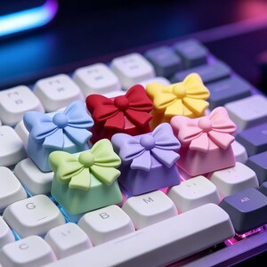 Könnte beinhalten: Ein Set aus sechs schleifenförmigen Tastenkappen auf einer mechanischen Tastatur. Die Tastenkappen sind hellblau, rot, gelb, hellgrün, lila und rosa. Jede Tastenkappe hat ein skulptiertes Schleifendesign auf der Oberseite.