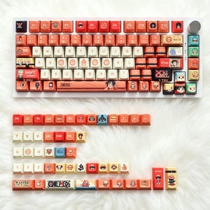 Puede incluir: Un teclado mecánico blanco con teclas personalizadas en tonos naranja, crema y azul. Las teclas muestran ilustraciones de personajes estilo anime, símbolos y el texto "ONE PIECE" y "WANTED". Se incluyen teclas sueltas adicionales.