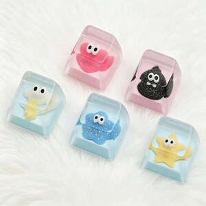 May include: Five custom mechanical keyboard keycaps featuring embedded cartoon characters with large white eyes. The set includes a pink, black, light blue, and yellow character design encased in clear resin, each with a unique glittery texture.
