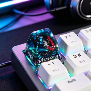 May include: A translucent, dark blue and black artisan keycap featuring a graphic of the character Venom with a long red tongue and the text VENOM printed in white. The keycap is installed on a mechanical keyboard with white keys and RGB lighting.