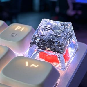 Puede incluir: Una tecla artesanal transparente con fragmentos negros, irregulares y de aspecto metálico incrustados en la resina. La tecla está situada en un teclado mecánico blanco junto a una tecla F1, con un interruptor rojo brillante visible debajo.