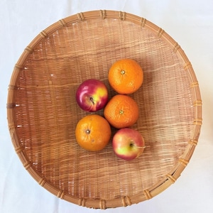May include: A large, round, woven bamboo basket with a reinforced rim, containing three bright orange citrus fruits and two smooth, round red and yellow apples arranged in the center of the shallow, light brown natural fiber container.