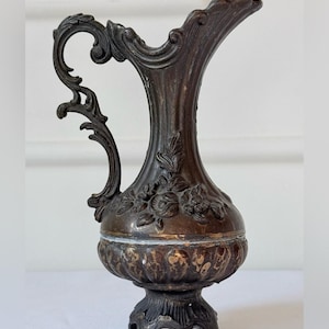 May include: A dark bronze-toned metal pitcher featuring an ornate, curved handle and a flared spout. The body is decorated with embossed floral motifs and a ribbed lower section, supported by a decorative base with three small, shaped feet.