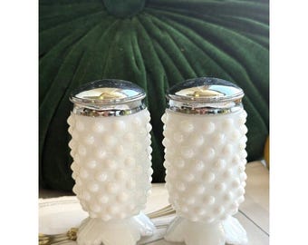 Puede incluir: Un par de saleros y pimenteros de vidrio lechoso blanco con textura de puntos en relieve y cuerpo cilíndrico. Cada pieza tiene una tapa de metal plateado pulido con acabado liso y redondeado, y una base acampanada con forma de pétalos.