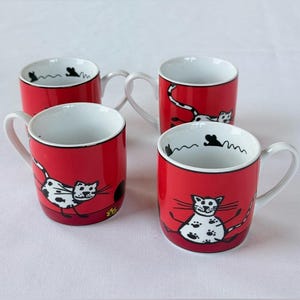 May include: A set of four red ceramic mugs featuring white interiors and black-and-white illustrations of cats. Each mug displays a cartoon cat with spots and a long tail, with small black mouse silhouettes depicted along the inner rim.