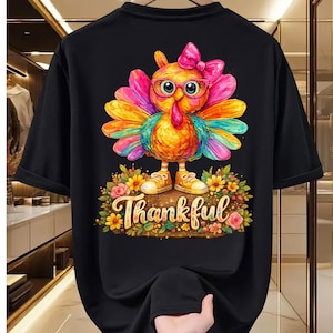 Op de afbeelding: Een zwart t-shirt met een kleurrijke afbeelding van een kalkoen met een roze bril en een roze strik. De kalkoen staat op een bloemenveld met het woord Thankful in een sierlijk lettertype eronder. Het ontwerp bevat levendige verfachtige streken.