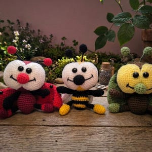 May include: Three crocheted stuffed toys featuring a red ladybird, a yellow and black striped bee, and a green beetle. Each toy has a white face, black button eyes, a stitched smile, and two antennae with small round tips on top of their heads.