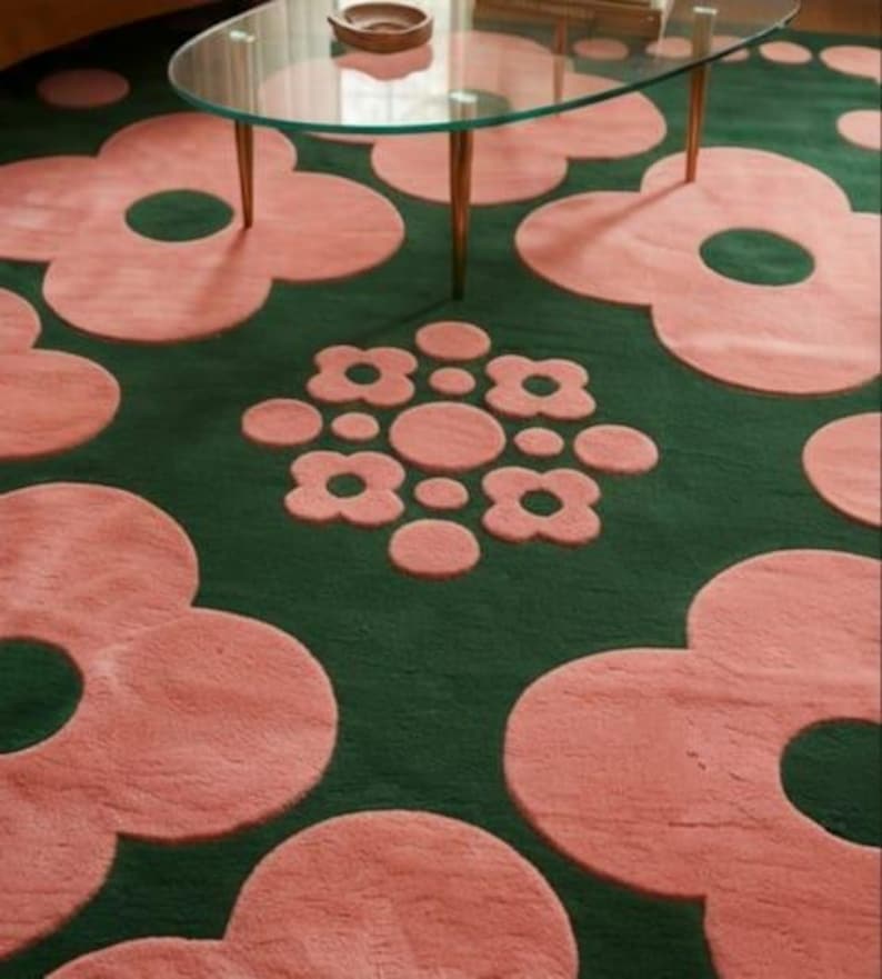 May include: A dark green area rug featuring a repeating pattern of large, soft pink flower shapes with circular centres. A central cluster of smaller pink flowers and circles is positioned between the larger floral motifs on the rug surface.