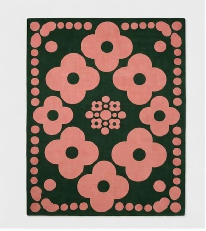 May include: A rectangular area rug featuring a dark forest green background with a repeating pattern of large, light pink four-petaled flower shapes. The border consists of a series of light pink circles and rounded shapes framing the central design.