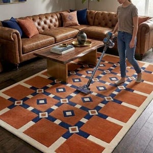 Könnte beinhalten: Ein rechteckiger Teppich mit einem geometrischen Muster aus großen rostorangen Quadraten, dunkelblauen sich kreuzenden Linien und kleinen cremefarbenen Quadraten. Der Teppich liegt auf einem dunklen Holzboden in einem Raum mit einem braunen Ledersofa.