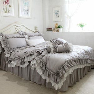 May include: A light gray bedding set featuring a duvet cover, pillow shams, and a bed skirt, all adorned with layered ruffles and white lace trim. The set includes a matching cylindrical bolster pillow and a rectangular white accent pillow.