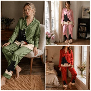 May include: A two-piece pyjama set consisting of a long-sleeved button-down shirt and matching trousers. The set is available in olive green, pink, or red satin fabric, each featuring a graphic print of a black cat sitting on a book.