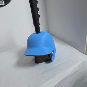 Pode incluir: Um capacete de beisebol azul claro montado num cabo preto com aderência texturizada. O capacete apresenta uma concha lisa e arredondada com uma pala frontal e proteção para as orelhas. O cabo preto estende-se verticalmente do capacete.