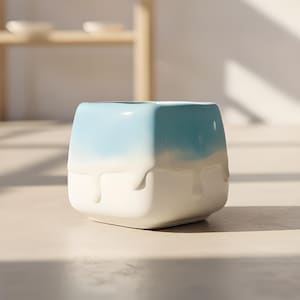 May include: A square ceramic planter featuring a smooth white base with a light blue gradient top. The design includes a decorative drip effect where the blue glaze appears to melt down over the white surface, creating a distinct layered appearance.