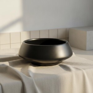 Pode incluir: Uma tigela preta fosca com um corpo largo e arredondado e uma base cônica. A tigela apresenta um acabamento liso e uniforme e um interior profundo e aberto. O design é caracterizado pela sua silhueta circular limpa e estrutura estável.