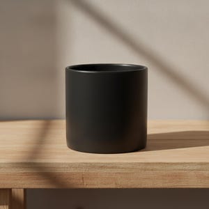Könnte beinhalten: Ein zylindrisches Gefäß mit einer glatten, mattschwarzen Oberfläche. Der Behälter zeichnet sich durch ein schlichtes Design mit geraden Seiten, einer weiten Öffnung an der Oberseite und einem flachen Boden aus, was ein einheitliches dunkles Erscheinungsbild ergibt.