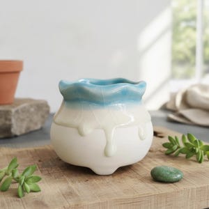 May include: A small ceramic planter with a rounded body and three short feet. The pot features a glossy finish with a blue rim that transitions into a cream-coloured glaze, creating a decorative drip effect over the matte white base.