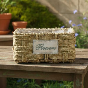 May include: A rectangular woven seagrass basket with a light tan finish. A rectangular fabric patch is centered on the front, featuring the word "Flowers" embroidered in a dark green script font with a decorative stitched border.