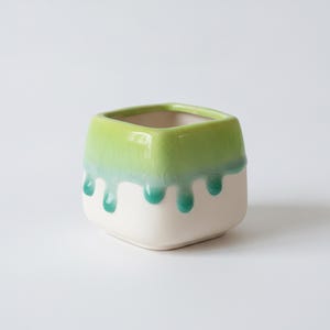 Puede incluir: Una maceta cuadrada de cerámica con una base blanca mate lisa y un esmalte verde lima brillante en la parte superior. El esmalte verde gotea por los lados en un patrón distintivo, creando un efecto de textura agrietada en la parte superior.