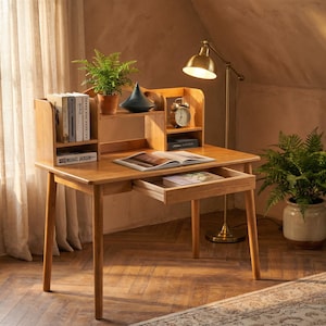 May include: A light brown wooden desk featuring a hutch with two open shelves, a single pull-out drawer, and four tapered legs. The desk surface holds an open book, while the hutch contains a potted fern, a black decorative object, and an analog clock.