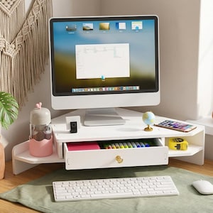 May include: A white corner monitor stand featuring a central drawer with a gold knob, side platforms for accessories, and a flat top surface. The stand holds a computer monitor, a small globe, a smartphone, and a yellow measuring tape.