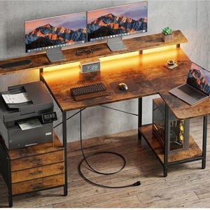 May include: An L-shaped computer desk with a rustic brown wood grain finish and a black metal frame. It features a raised monitor shelf with integrated LED lighting, three storage drawers, a printer stand, and a lower shelf for a computer tower.