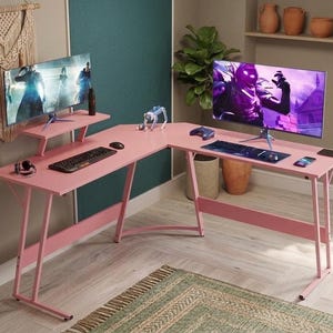 May include: An L-shaped pink gaming desk with a raised monitor shelf, thin metal legs, and a flat surface. The desk is set up with two computer monitors, a black keyboard, a computer mouse, a game controller, and a smartphone on the desktop.