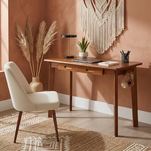 May include: A wooden desk with two drawers, featuring a laptop, a small potted plant, a black desk lamp, and a pen holder on its surface. A pair of headphones hangs from the side. An off-white upholstered chair with dark wooden legs sits nearby.