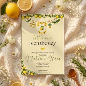 May include: A rectangular baby shower invitation card with a cream background featuring a watercolor illustration of a clothesline with baby shoes, a onesie, and a pacifier, decorated with lemons and white flowers, with the text little one is on the way.