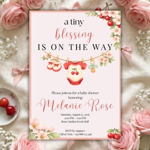 May include: A rectangular baby shower invitation card with a light pink background featuring a clothesline with baby booties, a cherry-print onesie, and a pacifier. The text reads: a tiny blessing IS ON THE WAY, honoring Melanie Rose, August 15, 2026.