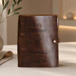 May include: A dark brown leather binder featuring the engraved text The Wedding Officiant Binder, a floral illustration, and the names Sarah & Anthony with the date 09.15.2026. The binder includes a side strap with a silver-toned metal snap closure.
