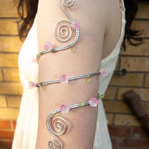 May include: A silver-toned wire arm cuff featuring a spiralling design at both ends. The metal wire is adorned with small, translucent pink flower-shaped beads and tiny green leaf-shaped accents distributed along the length of the coiled arm band.