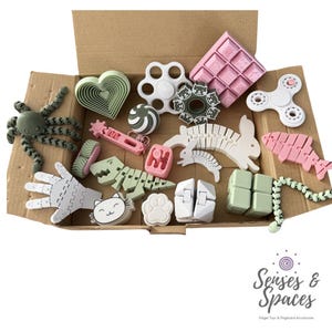 May include: A cardboard box filled with various sensory fidget toys in pastel and neutral colors, including an octopus, a heart, a white spinner, a pink waffle-shaped block, a dinosaur, a rabbit, a hand, and several geometric shapes and chains.