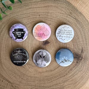 May include: Six circular pinback buttons featuring various designs: a purple cauldron with text, a pink watercolor mask and harp, a mountain graphic with text, a black button with soup kitchen text, a sword with wings, and a blue mountain peak graphic.