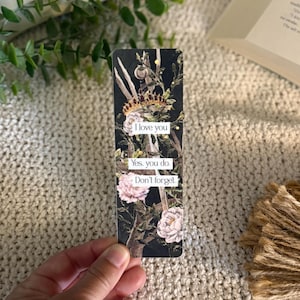 May include: A rectangular bookmark with a black background featuring a gold crown, green foliage, and light pink flowers. The bookmark displays the text: I love you. Yes, you do. Don't forget. The design is printed on a smooth, flat surface.