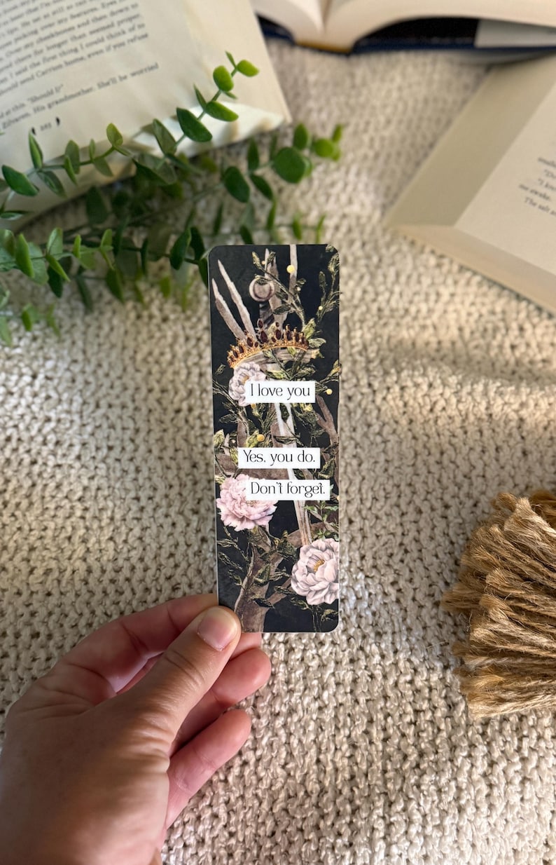 May include: A rectangular bookmark with a black background featuring a gold crown, green foliage, and light pink flowers. The bookmark displays the text: I love you. Yes, you do. Don't forget. The design is printed on a smooth, flat surface.