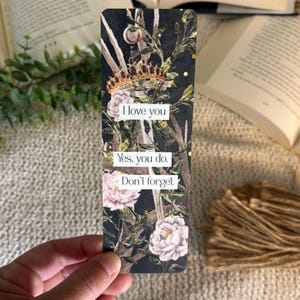May include: A rectangular bookmark featuring a dark background with a sword, a gold crown, and pink floral accents. The text reads: I love you. Yes, you do. Don't forget. The design includes intricate botanical elements and gold speckles throughout the surface.